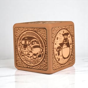 May include: A square, light brown wooden box with rounded edges. The box features detailed, engraved illustrations of characters from the animated film "My Neighbor Totoro" on its sides. The artwork includes Totoro and the Catbus.