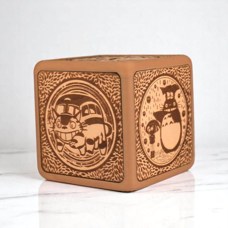May include: A square, light brown wooden box with rounded edges. The box features detailed, engraved illustrations of characters from the animated film "My Neighbor Totoro" on its sides. The artwork includes Totoro and the Catbus.
