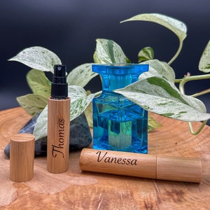 May include: Two personalized bamboo roller bottles with black spray tops. The bottle on the left is labeled "Thomas" and the bottle on the right is labeled "Vanessa". A blue glass bottle is in the background.