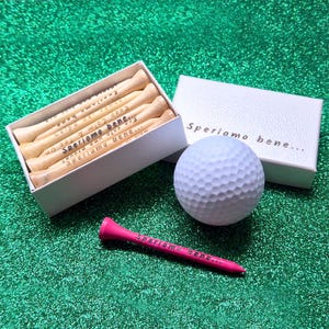 Puede incluir: Un juego de regalo de golf con una pelota blanca, una caja de tees de madera y un tee rosa. La caja y el tee rosa están inscritos con "Speriamo bene...". Los tees de la caja también tienen texto.