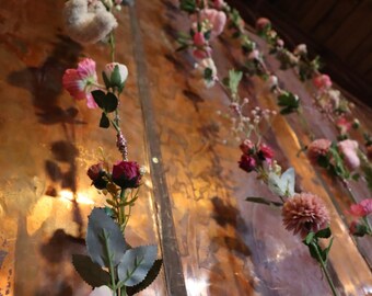 May include: A display of artificial flowers in shades of pink, red, and white against a textured, copper-colored background. The flowers are arranged in a cascading pattern, creating a decorative and visually appealing arrangement.