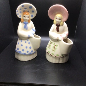 May include: Two vintage ceramic figurines of women wearing hats and dresses. The woman on the left is wearing a blue and white polka dot dress and a blue hat with white polka dots. The woman on the right is wearing a green and white dress with a pink hat. Both figurines have a white base and are holding a white vase.