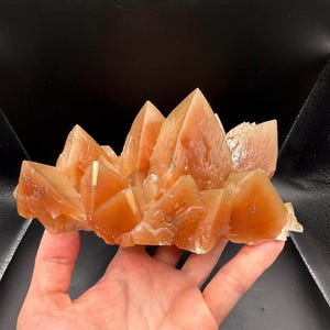 May include: A cluster of translucent, orange-hued calcite crystals featuring sharp, pointed terminations and a jagged, multi-faceted structure. The mineral specimen displays varying shades of amber and burnt orange with visible internal crystalline layers.
