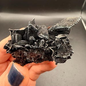 May include: A raw mineral specimen featuring a cluster of dark, metallic, and crystalline structures with sharp, angular edges. The base is composed of a porous, dark brown, earthy material that supports the glossy, charcoal-colored mineral formations.