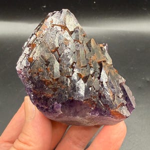 May include: A cluster of deep purple amethyst crystals featuring sharp, pointed terminations and prominent reddish-brown iron oxide inclusions embedded within the crystalline structure, forming a dense, jagged, and multifaceted mineral specimen.