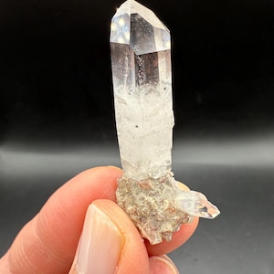May include: A clear quartz crystal with a pointed tip and a rough, textured base. The crystal is transparent, allowing light to pass through, and is held against a dark background. The base has a mix of beige and gray tones.
