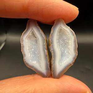 May include: Two matching agate slices, each with a light blue interior and a white crystalline center. The outer edges are a mix of brown and gray. The slices are held between fingers against a dark background.