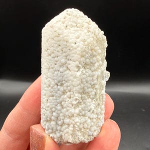 May include: A white, cylindrical mineral specimen with a textured, bubbly surface. The mineral has a rounded top and is held against a black background. The surface is covered in small, pearl-like formations.