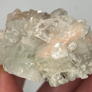 May include: A cluster of clear, translucent quartz crystals with a pink, translucent, elongated crystal in the center. The crystals are arranged in a cluster, with the pink crystal in the center.
