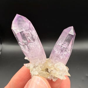 May include: A cluster of amethyst crystals with a pale purple hue. Two large, pointed crystals rise from a base of smaller, light-colored crystals. The crystals have a translucent quality, with visible internal structures.