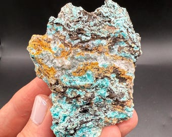 May include: A rough-hewn mineral specimen with a striking combination of colors. The rock features layers of turquoise, brown, and gold hues, with a crystalline white band running through the center. The specimen is held against a dark background.