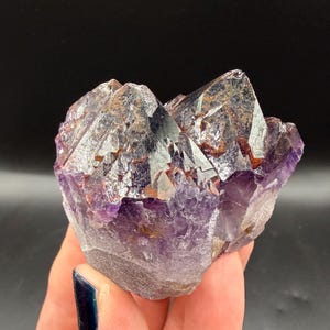 May include: A raw amethyst crystal cluster featuring deep purple hues and translucent, jagged points. The mineral specimen displays natural facets with dark, earthy inclusions embedded within the crystalline structure, creating a textured appearance.