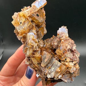 May include: A cluster of crystalline minerals with a mix of amber, brown, and clear hues. The crystals have a layered structure and are interspersed with small, dark inclusions. The overall shape is irregular and textured, with a natural, earthy appearance.