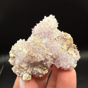 May include: A cluster of light purple crystals with a crystalline structure. The crystals are clustered together, with some areas appearing more dense than others. The base of the cluster is a light tan color, with some darker brown spots.