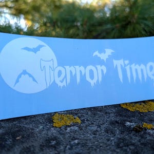 Pode incluir: Um autocolante retangular azul-claro com um gráfico branco de uma lua cheia, três morcegos a voar e o texto Terror Time numa fonte estilizada e a escorrer, impresso numa superfície plana com um acabamento mate.