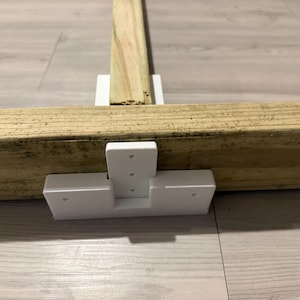 May include: A white T-shaped plastic bracket connecting two light brown wooden beams. The bracket features a vertical section with three small circular holes and a horizontal base with two small circular holes on each side, securing the wood pieces.