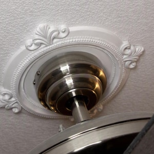 May include: A brushed nickel ceiling fan motor housing mounted to a white ceiling. The fan is surrounded by a circular white ceiling medallion featuring a decorative rope border and four symmetrical leaf-like carvings at the cardinal points.