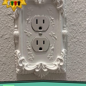 May include: A white electrical outlet wall plate featuring an ornate, raised relief frame with floral and scrollwork details. The plate includes a central screw and a starburst graphic in the corner with the text Matching Screws Included.