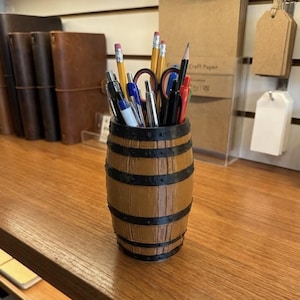Puede incluir: Un organizador de escritorio con forma de barril de madera, lleno de bolígrafos, lápices y tijeras. El organizador es marrón con bandas negras y se encuentra sobre una superficie de madera. Otros artículos de papelería son visibles en el fondo.