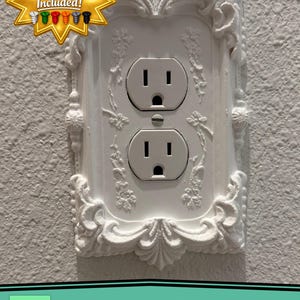 May include: A white electrical outlet cover plate featuring an ornate, raised relief border with floral and scrollwork details. The plate includes a central opening for a standard duplex receptacle and text stating Matching Screws Included.