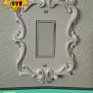 May include: A white rectangular light switch cover plate featuring an ornate, raised scrollwork border. The plate is secured with two flat-head screws and surrounds a single white rocker-style light switch. A star-shaped graphic indicates matching screws are included.