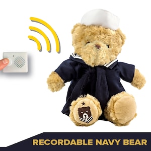 Pode incluir: Um urso de pelúcia vestido com um uniforme de marinheiro azul marinho e um chapéu branco. O urso tem pelo castanho claro e um pequeno emblema no pé. Um pequeno dispositivo branco com um botão é mostrado ao lado do urso, com ondas sonoras amarelas.