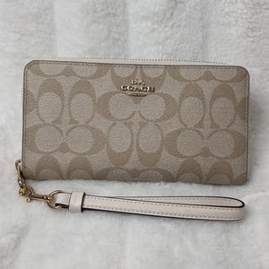 May include: A beige Coach wallet with a gold zipper and a white wrist strap. The wallet has a signature Coach pattern.