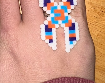 Peut inclure: Une bague colorée faite de perles en plastique fusionnées. La bague présente un motif géométrique avec des perles blanches, bleues, oranges et violettes. La bague est portée sur un doigt, mettant en valeur le savoir-faire artisanal.