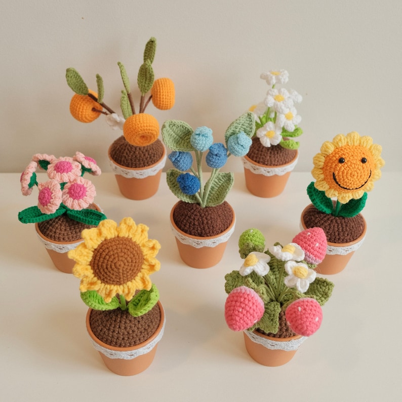 May include: A collection of seven crocheted potted plants. The plants include a sunflower, a daisy, a blueberry bush, a strawberry plant, a citrus tree, and two flower arrangements. The pots are all white with a lace trim.