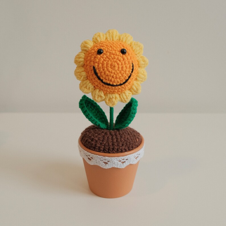 May include: A crocheted sunflower with a smiley face, in a brown pot with a white lace trim. The sunflower is yellow and has green leaves.
