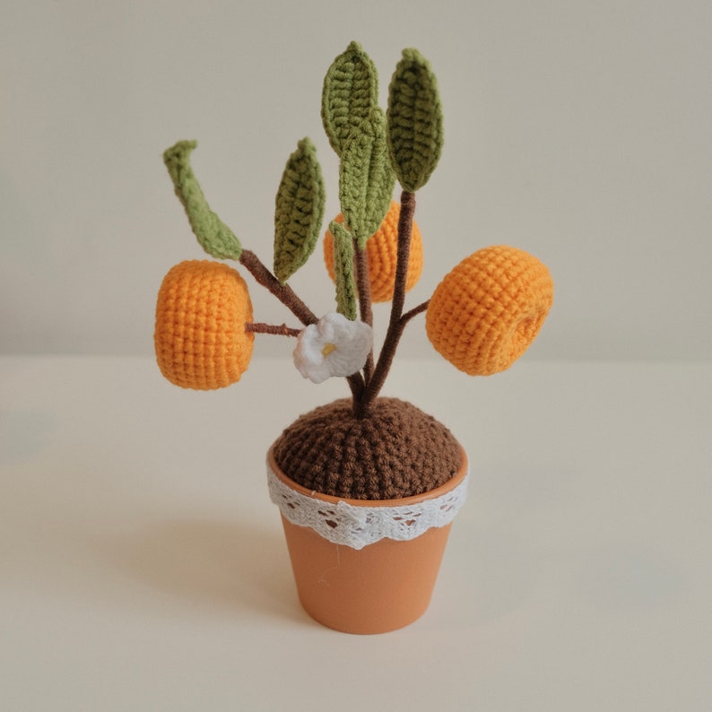 May include: A crocheted orange tree with three orange fruits and a white flower. The tree is potted in a brown pot with a white lace trim.