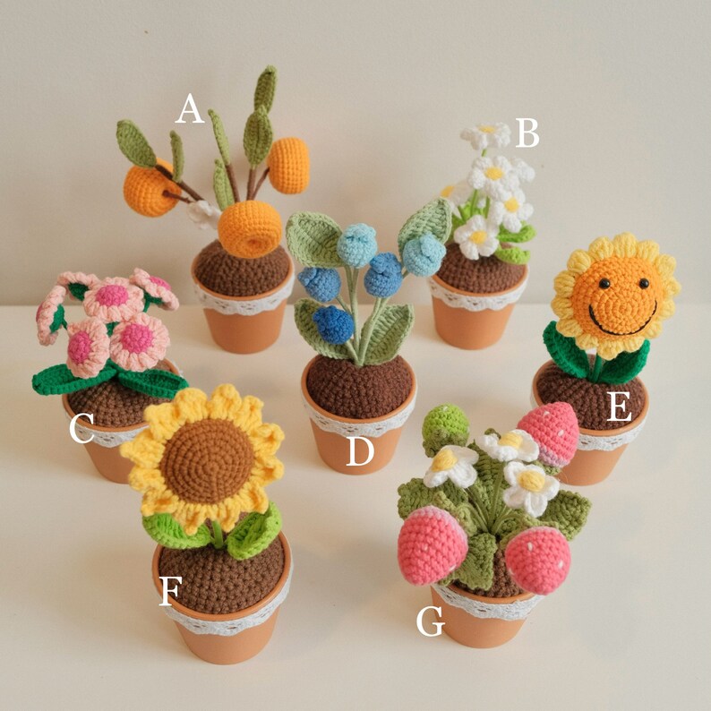 May include: A collection of 7 crocheted potted plants. The plants include a sunflower, a daisy, a strawberry, a blueberry bush, a pink flower, an orange tree, and a smiling sunflower. Each plant is in a small brown pot with white lace trim.