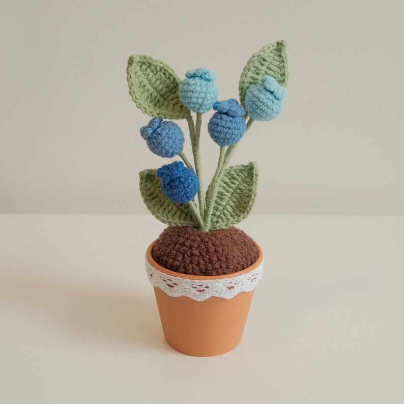 May include: A crocheted blueberry plant in a brown pot with white lace trim. The plant has green leaves and blue and light blue berries.