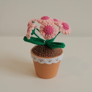 May include: A crocheted flower pot with pink flowers and green leaves. The pot is brown with a white lace trim.