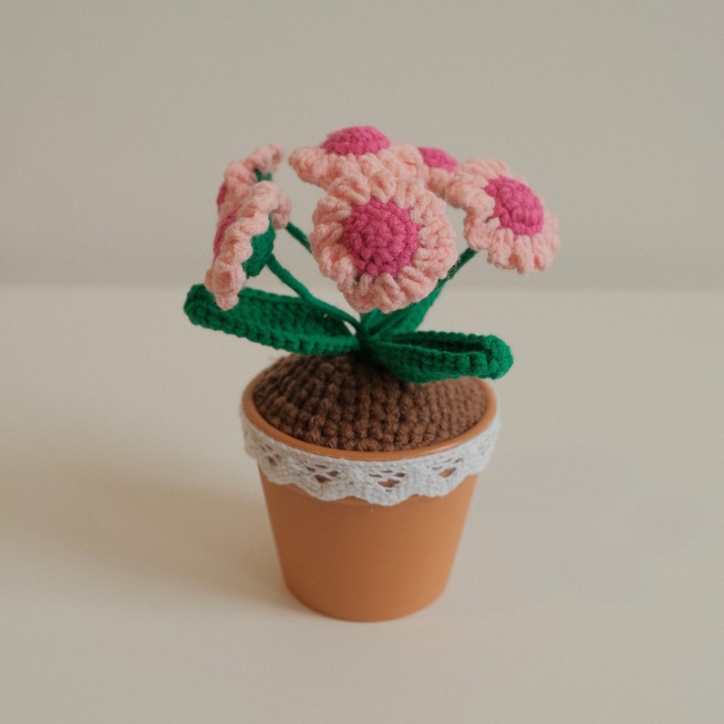 May include: A crocheted flower pot with pink flowers and green leaves. The pot is brown with a white lace trim.