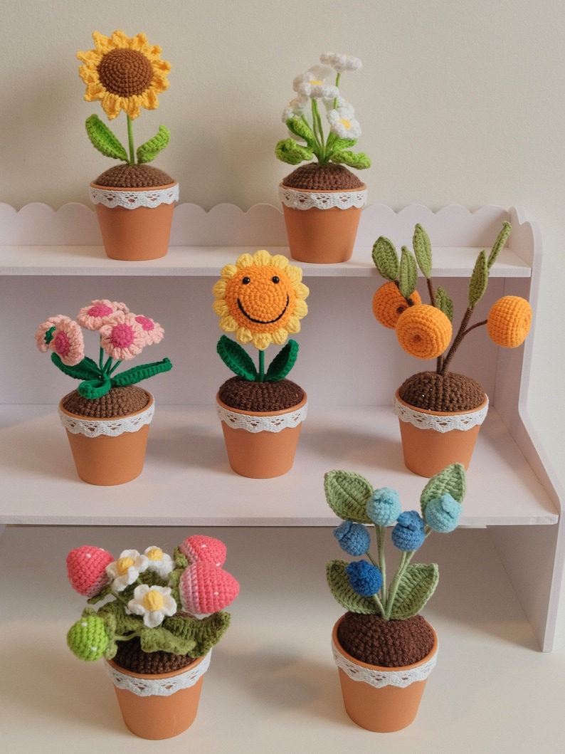 May include: A set of seven crocheted potted plants, each with a different flower or fruit. The plants are arranged on a white shelf with scalloped edges. The flowers include a sunflower, daisies, a pink flower, a smiley face flower, and strawberries. The fruits include oranges and blueberries.