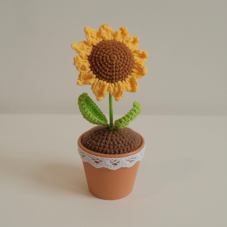 May include: A crocheted sunflower with yellow petals and a brown center, in a brown pot with a white lace trim.