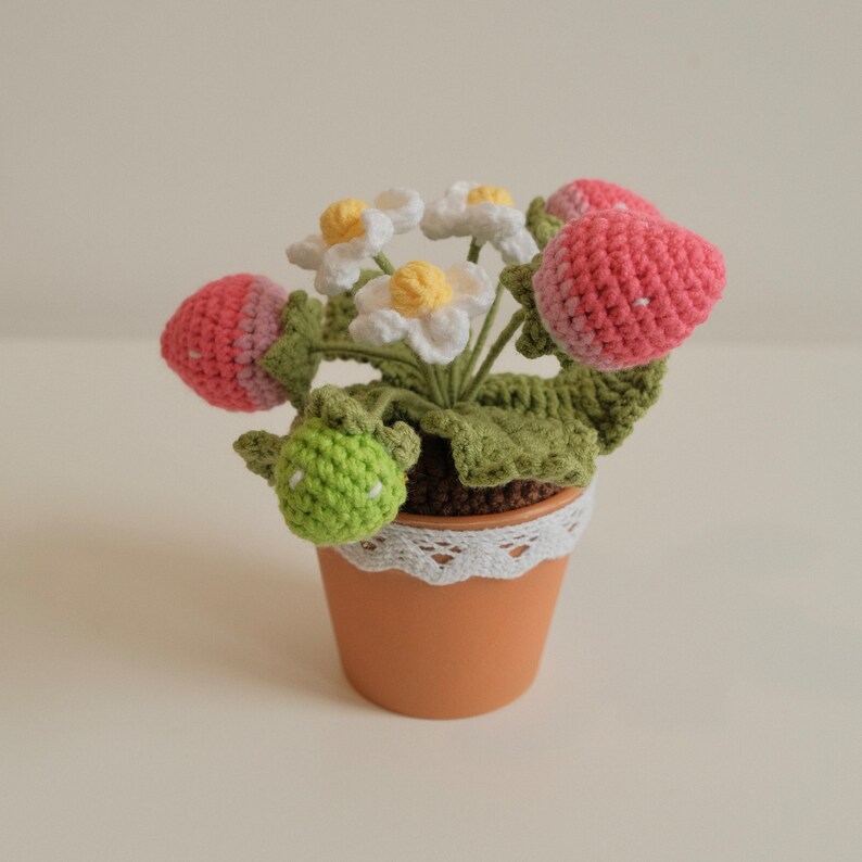 May include: A crocheted strawberry plant in a brown pot with white lace trim. The plant has pink and white strawberries and white flowers.