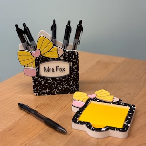 May include: A composition notebook-themed pen holder with black pens, a matching notepad, and a single pen on a wooden surface. The pen holder and notepad feature a yellow bow and the name "Mrs. Fox".
