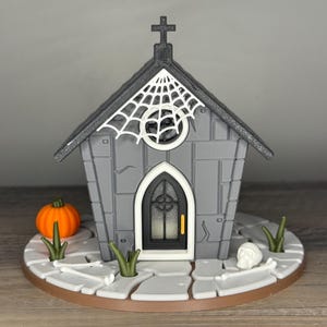 May include: A gray miniature Halloween church with a black roof, a white spiderweb, and a cross. An orange pumpkin, skull, and green grass accents the base. The church has a black door and a round window.