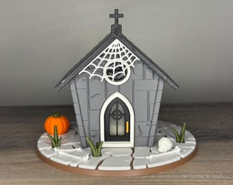 May include: A gray miniature Halloween church with a black roof, a white spiderweb, and a cross. An orange pumpkin, skull, and green grass accents the base. The church has a black door and a round window.
