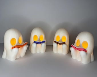 May include: Four white ghost figurines with glowing eyes, each holding a different colored book in front of their faces. The ghosts are arranged in a row on a white surface.