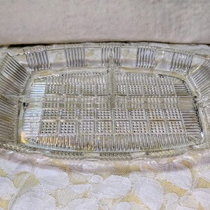 May include: A clear, rectangular glass serving tray with four divided sections. The tray features a textured, grid-like pattern in the center and vertical lines along the edges. It has curved handles on each end, suitable for serving appetizers or snacks.