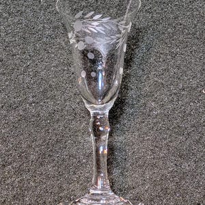 May include: A clear, stemmed glass with etched floral and dot designs. The glass has a flared rim and a slender stem, suitable for serving wine or other beverages. The glass is likely made of crystal or a similar material.