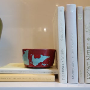 May include: A bookshelf with a variety of books, including "The Princess Within" by Serita Ann Jakes, "Mommy Wars" by Leslie Morgan Steiner, "Second Nature" by Alice Hoffman, and "The American Way of Strategy". There is a small red and blue ceramic bowl on the shelf.