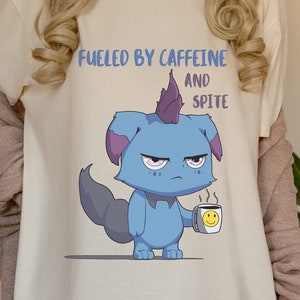 May include: Cream-colored t-shirt with the text "Fueled by Caffeine and Spite" above a cartoon blue creature holding a coffee mug with a smiley face. The creature has a purple mohawk and a grumpy expression.