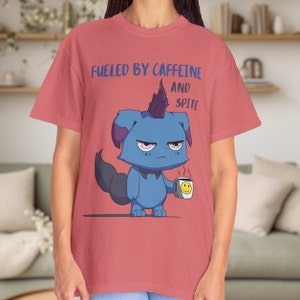 May include: A coral-colored t-shirt featuring the text "Fueled by Caffeine and Spite" above a cartoon character. The blue character holds a coffee cup with a smiley face. The character has a grumpy expression.