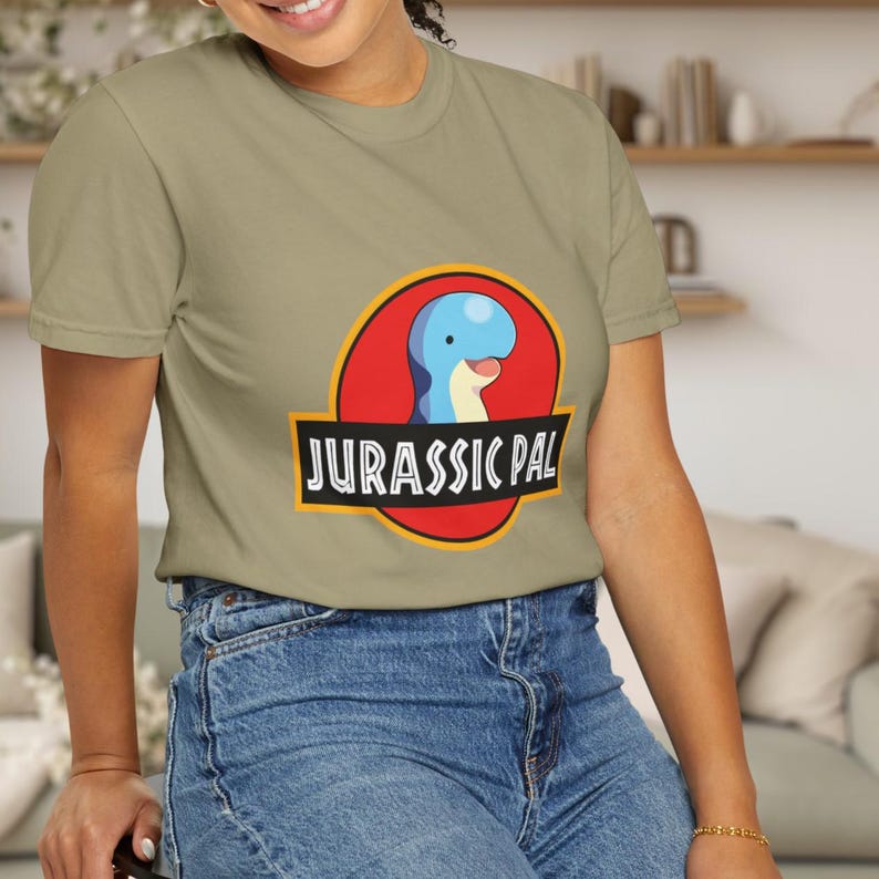 May include: A sage green short-sleeve t-shirt featuring a graphic of a blue dinosaur head inside a red circle with a yellow border. Below the circle, a black banner contains the white text JURASSIC PAL in a bold, sans-serif font style.