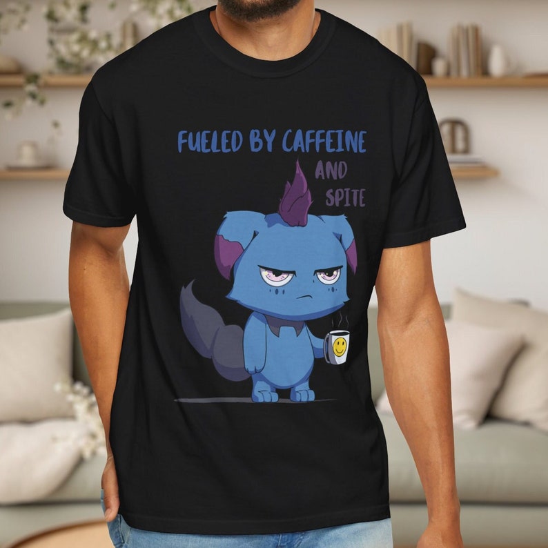 May include: Black t-shirt featuring a grumpy blue cartoon creature holding a coffee cup with a smiley face. The shirt has the text "Fueled by Caffeine and Spite" in blue lettering. The design is centered on the front of the shirt.