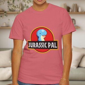 May include: A dusty rose short-sleeved t-shirt featuring a graphic of a blue dinosaur head inside a red circle with a yellow border. Below the circle, a black rectangular banner contains the white text JURASSIC PAL in a bold, sans-serif font.